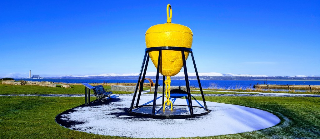 A large yellow buoy sculpture on a circular black base on the Bo'ness Foreshore, with a scenic view of the River Forth and hills in the background under a clear blue sky.