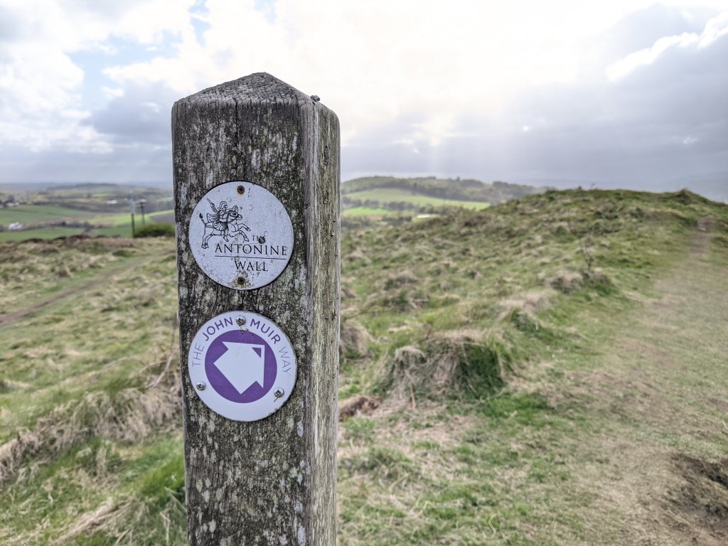 A wooden signpost indicating the Antonine Wall and the John Muir Way, set against a backdrop of rolling hills and cloudy skies.