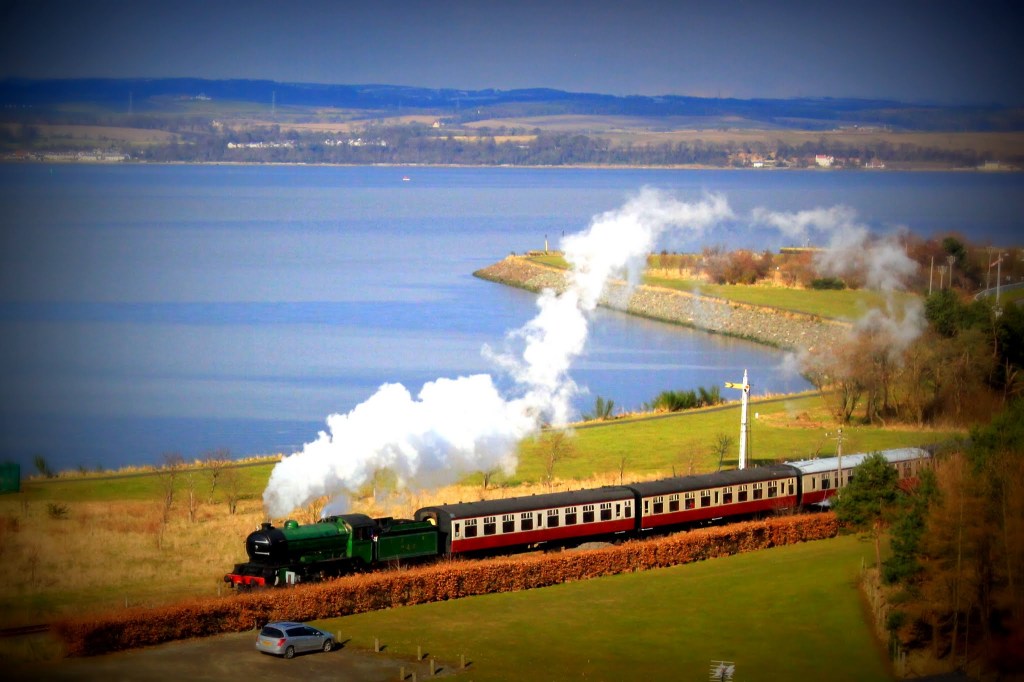 A steam locomotive pulling a vintage train along a scenic waterfront, with smoke billowing from its chimney and a backdrop of rolling hills and a blue sky.
