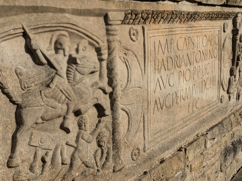 Close-up of a carved stone with Roman inscriptions and figures, depicting soldiers and symbols related to the Antonine Wall.